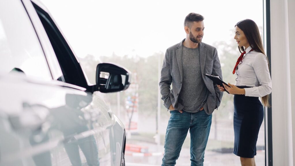 Una asesora conversa con un cliente junto a un coche, en una sala luminosa